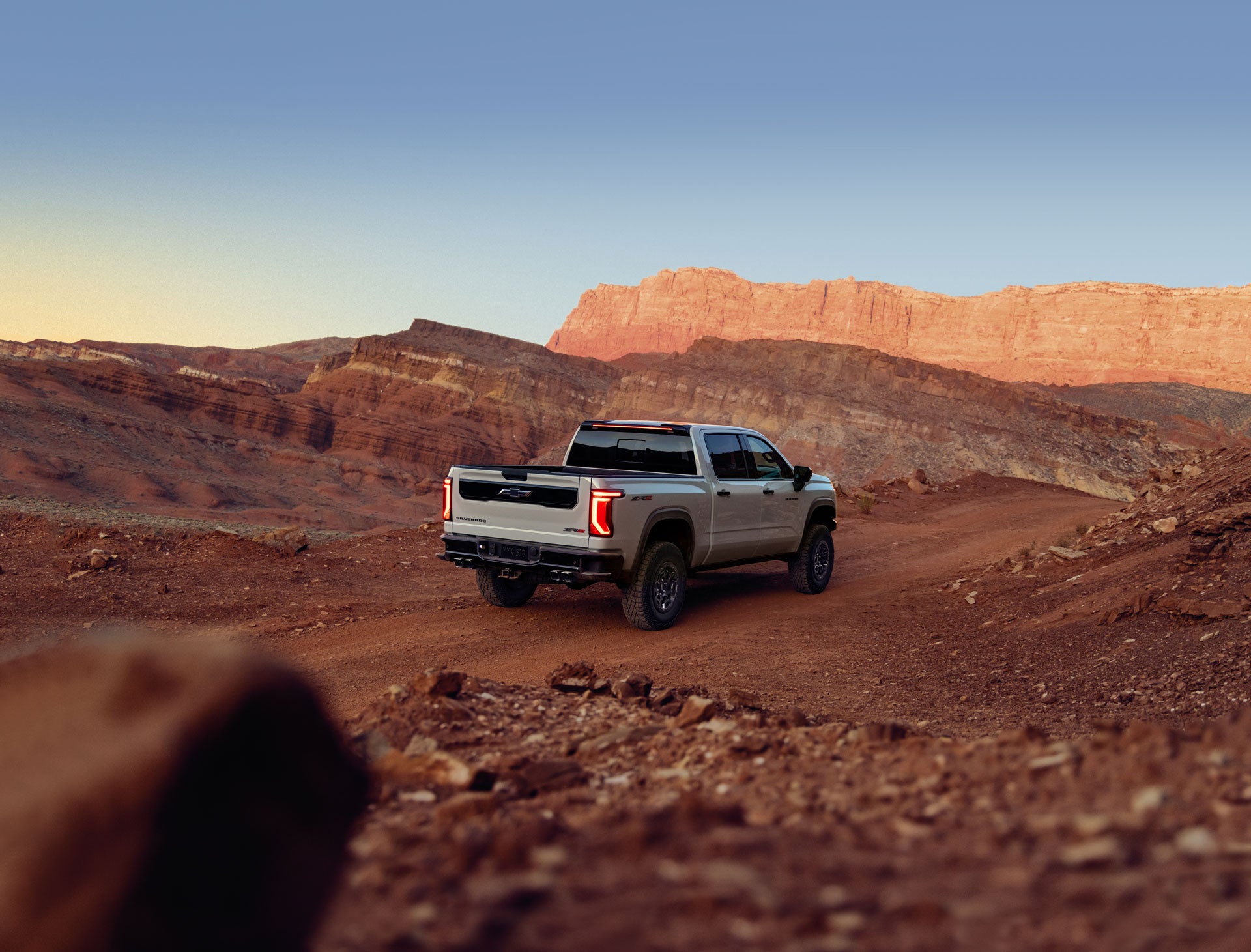 Rear view of a gray 2027 Chevrolet Silverado 1500 on a dirt road in a desert landscape during golden hour.
